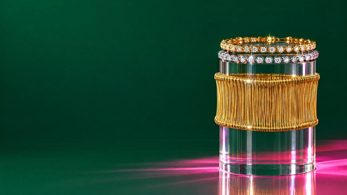 Bracciali tennis argento e oro e bracciale rigido dorato impilati su un cilindro trasparente, fondo verde smeraldo